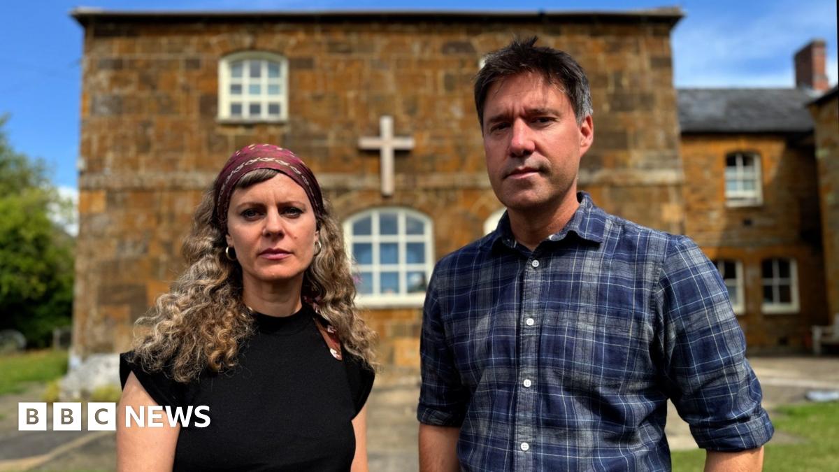 Ellena Wood with long curly hair, a head scarf and a black top stands beside Jon Ironmonger, who is wearing a blue-checked shirt in front of a small orange-brick chapel building.