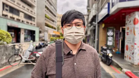 BBC / Tessa Wong A man with salt and pepper hair wearing wire-rimmed glasses and a face mask poses for the camera. He is wearing a brown polo shirt and you can see his bag strap on his shoulder. In the background you can see the streets of Taipei including parked bicycles and motorcycles.