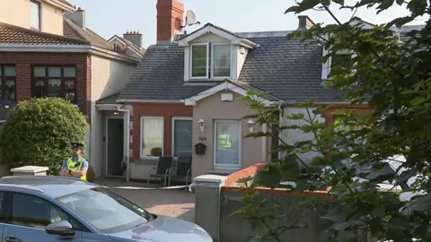 RTE Officer stands outside a house. A car is parked. Some chairs are sitting outside the house. 