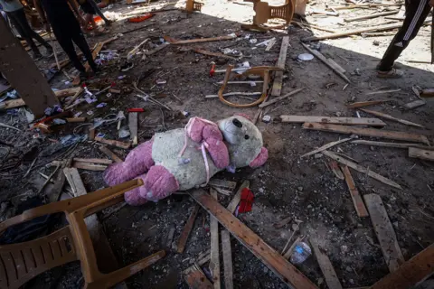 Anadolu via Getty Images A muddy pink and white teddy bear lies among debris of wood beams, broken chairs and playing cards as people can be seen standing in the background of the image, at the site of the blast on Monday.