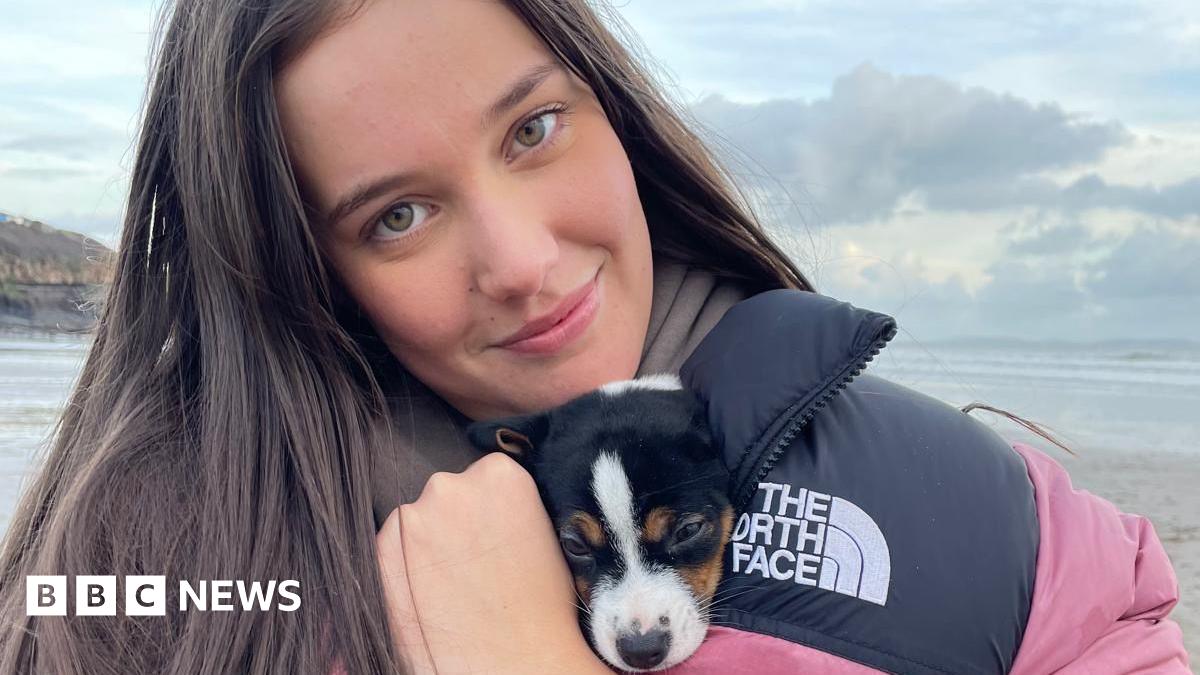 Photograph of 18-year-old Sally Allen on a beach. She has long dark straight hair, hazel eyes and looks at the camera with a smile. She holds a small puppy inside the collar of her north face puffer coat.