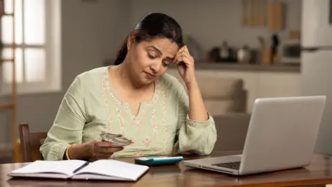 Getty Images Middle-aged woman with money and calculator in front of an open laptop on a desk