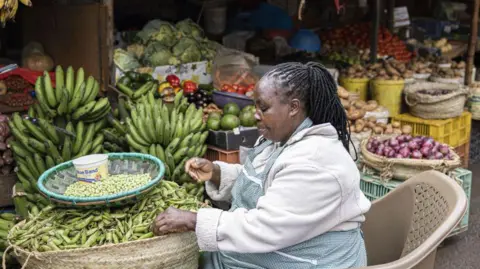 Bloomberg via Getty Images A woman, a vendor, at a market stall in Kenya with green peas, green bananas and other groceries