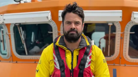 Simon Jones/BBC Dan Sinclair - a man with black hair, a black beard and a black moustache. He is  wearing a yellow RNLI wetsuit and a red life jacket. The picture has been taken from a boat in the sea.