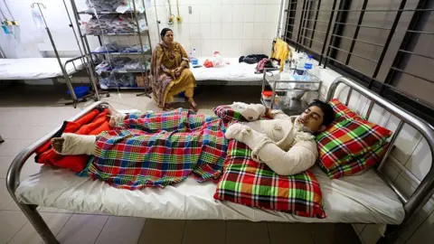 Getty Images A boy covered in bandages, lying on a hospital bed. A woman in a yellow dress sits on the bed beside him and watches him.