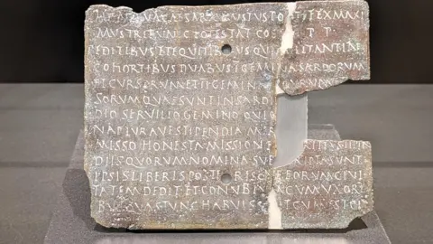 Thea Sommerschield A square stone coloured plaque with a big chunk on its right hand side missing