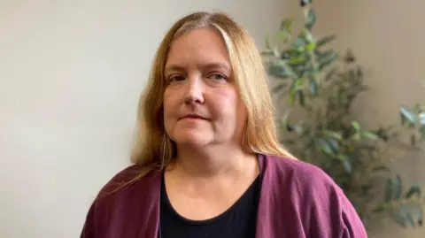 BBC Anna is a middle-aged woman with long hair. She's wearing a black top and a purple cardigan. Behind her are the green leaves of some indoor plant.
