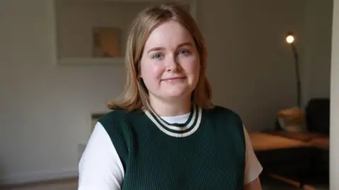 Bethan Williams smiles at the camera. She is sat in her front room and there is a lamp on in the background 