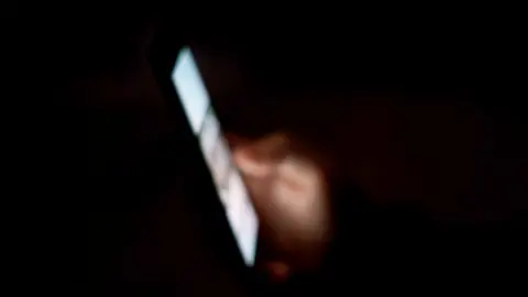 A smartphone display is lit up in a darkened room with the blurred image just visible of the hand holding it.