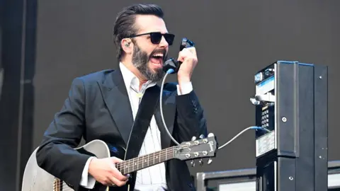 Getty Images Ben Schneider singing into a payphone handset as a prop on stage, also holding a guitar