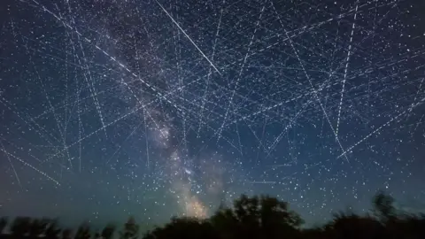 Getty Images An image of the night sky taken on a long exposure which shows it criss-crossed with dotted streaks from satellites moving through the sky. At the bottom of the image a dark line of trees is silhouetted while you can also see the stars and bright light  of the milky way in the image.