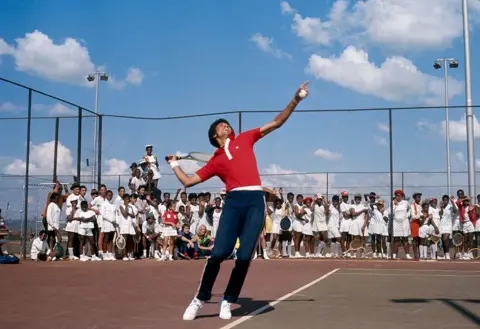 Gerry Cranham / Offside Arthur Ashe in red shirt and navy blue tracksuit bottoms serves as a crowd of South African children in tennis whites watch him from behind a tennis court fence in Soweto - November 1973.