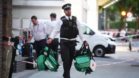 PA Media A police officer carries green first aid bags away from the scene of the stabbings.  Police tape blocking the road is visible behind him. 