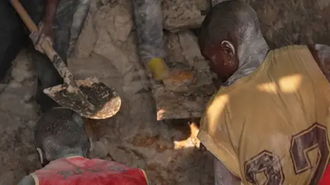 Hassan Lali / BBC Two miners with their backs to the camera bend over as they dig in a pit at Rubaya mine in eastern DR Congo. The one in the left wears a red tank top and his colleague on the right wears a yellow sports shirt with numbers printed on the back. In the background the metal spade of another miner can be seen.