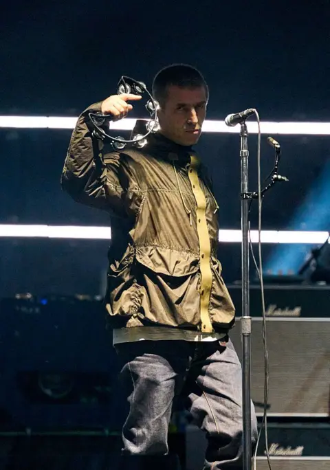 Getty Images Liam Gallagher is pictured standing in front of a microphone on stage while holding a tambourine. He is wearing a green jacket.