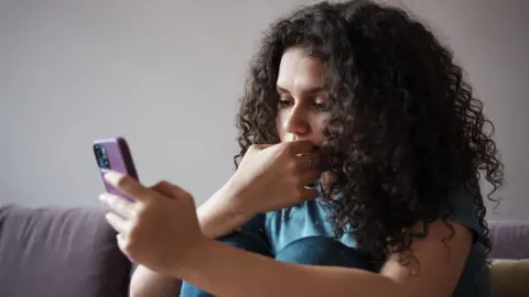 Getty Images A young woman with dark curly hair looks at a smartphone screen with a concerned face