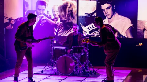 Graham Turner Three men on stage playing instruments as pictures of Elvis appear in the background
