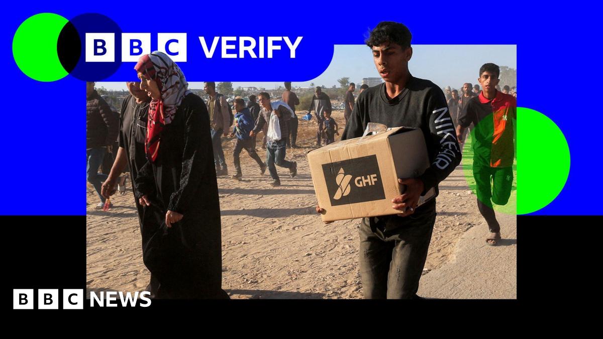 An image showing a young man carrying an box of aid with the GHF logo emblazoned on it. He is wearing a dark shirt with Nike Air written on it. Several other people are walking alongside him, and the image is imposed over the BBC Verify branding and colours.