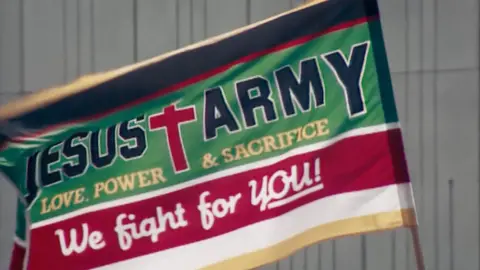 BBC/Docsville Studios The Jesus Army flag in green and red with a large red cross, reads 'Love, Power & Sacrifice; We right for YOU!'