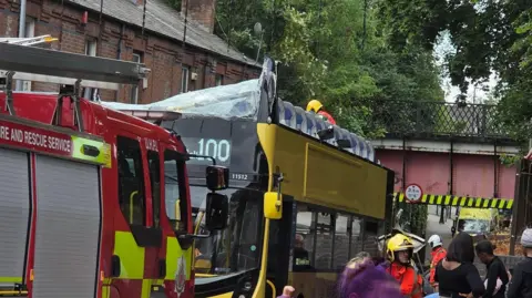 X A yellow double-decker bus has a smashed top windscreen and no roof, leaving the top floor seats exposed. A fire engine is parked in front of it and a man in a fireman's helmet can be seen stand on the street next to it speaking to people. 