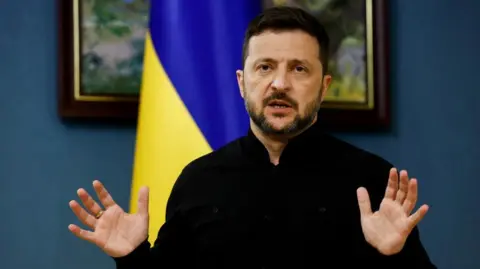 Reuters Ukraine's president gestures with his hands in a dark top with a Ukrainian blue and yellow flag behind him