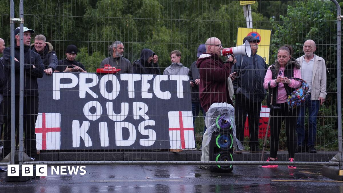 A group of about 12 people stand behind a fence as part of a protest. A large sign reads 'Protect our kids'. One of the men has a megaphone to his mouth and a stack of speakers by his feet