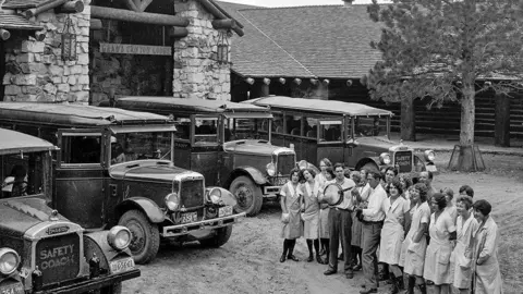National Park Service A black-and-white photo of old-fashioned buses in front of a stone building, with a line of women in cleaning uniforms and two men playing stringed instruments