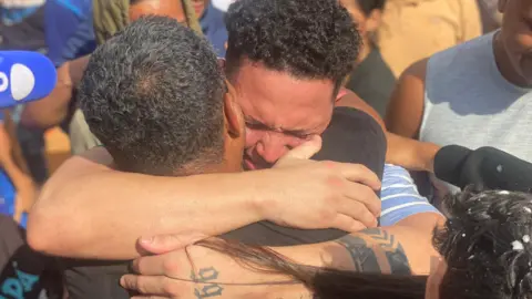 Gustavo Ocando Mervin Yamarte cries as he hugs a relative in the Los Pescadores neighbourhood in Maracaibo. On his arms, he has several tattoos. A microphone can be seen in one corner as journalists try to capture the moment of the reunion