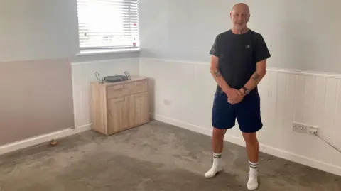 Dave Hemmings, who is bald and has a short beard, stands in his empty flat. There is only a carpet and a small cupboard left in the bare space. 