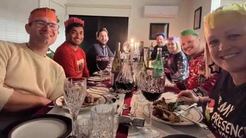Joshua Robb A group of people sit around a dinner wearing Christmas themed outfits and paper crowns, eating a Christmas meal