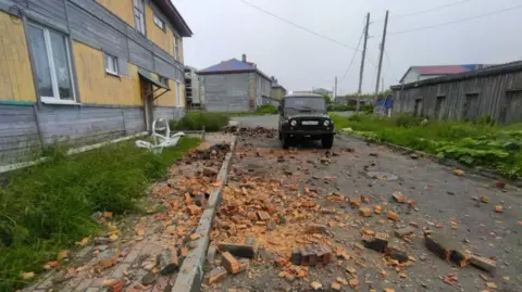 S. Lakamov/Anadolu/Getty Images A 4x4 truck is parked in a street with broken bricks and rubble all around it. To the left is a yellow wooden building with a grass verge. 
The sky appears overcast, and other buildings can be seen in the background. Telephone wires criss-cross behind the truck
