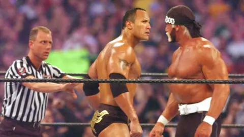 Getty Images Dwayne Johnson and Hulk Hogan square up, eyeing each other menacingly, dwarfing the referee who's trying to keep order next to them, in the ring at Wrestlemaia X8 in 2002