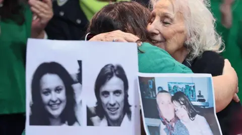 EPA A black-and-white composite photo of the missing couple can be seen in front of Adriana Metz. A smaller photo shows Adriana as a child with her grandmother 