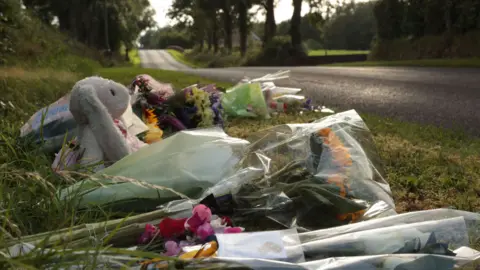 PA Media A teddy and flowers are lying on the side of a country road.