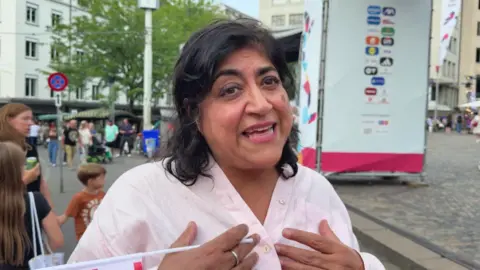 Director Gurinder Chadha wears a pink shirt and holds a mini England flag as she speaks 