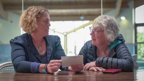 BBC News Two women sat at a table next to each other, looking at family photographs. They look at each other as they hold the photos. 
The woman on the left is wearing a navy jacket and has short ginger curly hair. The women on the right has short grey hair, glasses and is wearing a dark coat. 