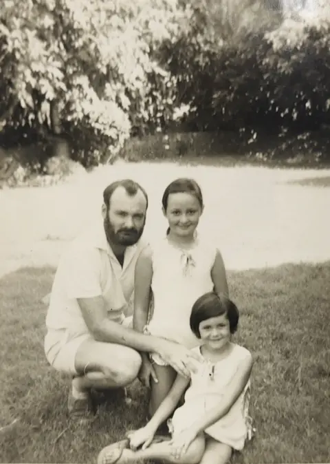 Michele Dougherty Ten year-old Michele stands in the centre of a black and white picture from 1972 with her father to the right and her younger sister in front. They appear to be in a sunny garden and wearing white or bright short sleeves.