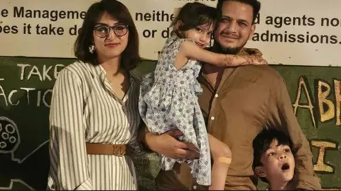 Javid Ali, hair short and wearing a brown button-down shirt, stands holding his daughter Amani, wearing a dress, beside his wife Maryam Ali, wearing a belted dress, glasses, and her dark hair short. Their son Zayn stands in front of them, Javid's arm around him.
