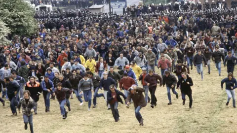 Getty Images Dozens, possibly hundreds, of men run across a large patch of grass. Police officers, some of them on horses, can be seen in the background.