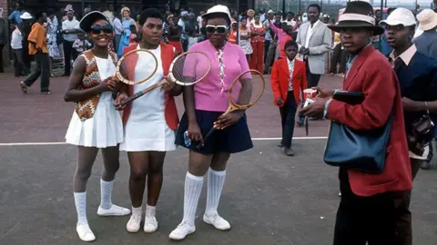 Gerry Cranham / Offside Young girls, some in in tennis whites and sunglasses, pose with racquets as boys in suits and hats walk by. They are part of crowd gathered in Soweto to see Arthur Ashe in November 1973.
