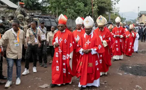 Reuters Catholic bishops in red robes walk along a dirt street near Goma's cathedral after celebrating the mass following the beatification of Floribert Bwana Chui Bin Kositi. A crowd of top officials in suits follow them. Behind the cleric to the left can be seen a vehicle containing uniformed M23 rebel soldiers.