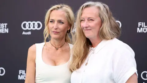Getty Images Actress Gillian Anderson on the left, wearing a sleeveless white dress with her blonde hair in a ponytail, looks away from the camera. She is stood next to author Raynor Winn, who has long and wavy fair hair and wears a short sleeved white shirt.