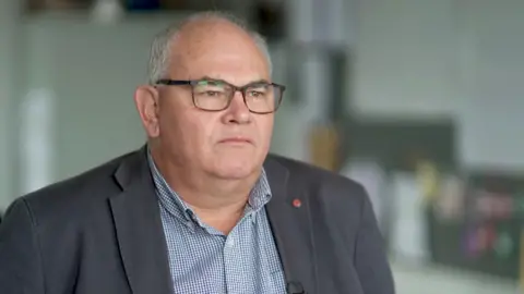 Paul Abbott looks from left to right - he is wearing glasses, a dark blazer, and a checkered shirt. The background is an indoor setting with indistinct objects and furniture.
