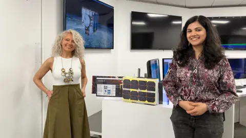 Emily Coady-Stemp/BBC Two women stand either side of a small model satellite with black panels on the side and painted yellow. They are standing in front of computer and TV screens, one showing an image of a satellite in space.