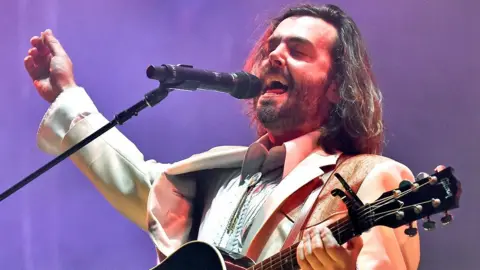 Getty Images Lord Huron singer Ben Schneider on stage in a white suit with mouth open and eyes closed mid song, with one hand on a guitar neck and the other in the air. At the 2022 Bonnaroo Music & Arts festival in Tennessee.
