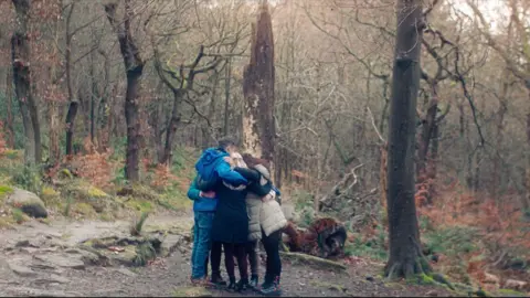Docsville Studios A group of people in rain jackets give each other a group hug in a wooded area.