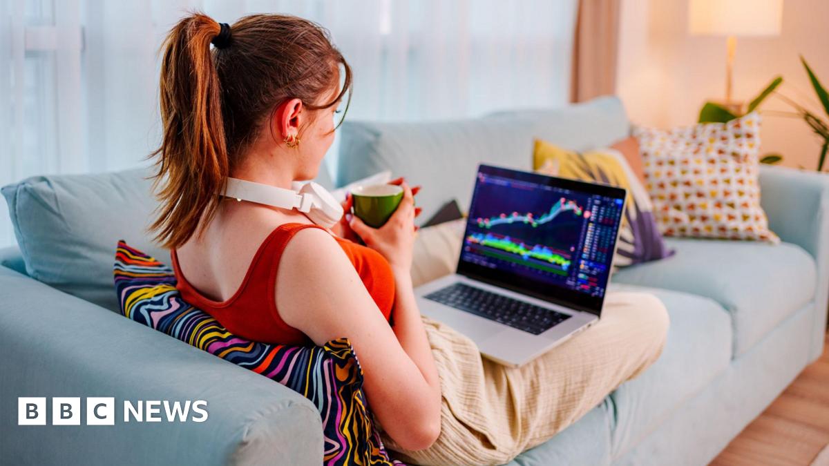 Young woman sits on the couch with her feet up, coffee cup in hand, checking performance of shares on a laptop.