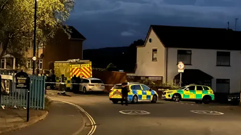Two police cars and an ambulance can be seen parked on a residential street. The ambulance is behind a strip of police tape cordoning off the road. It is dark and there are no lights from any of the houses visible  