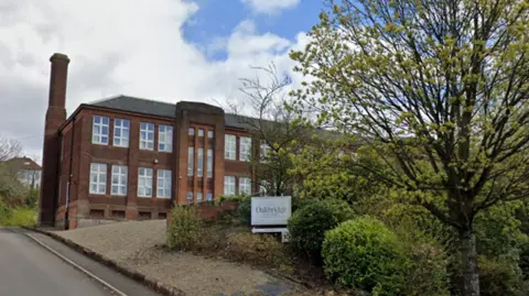 Google A road leading towards a red brick building with several large trees in front of it. A sign saying Oakbridge is in front of it. 