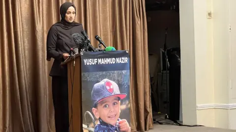 Jamie Coulson/BBC A woman with her hair covered stands in front of a lectern with the picture of a young boy smiling on it. It has the words Yusuf Mahmud Nazir at the top of the lectern above the image.